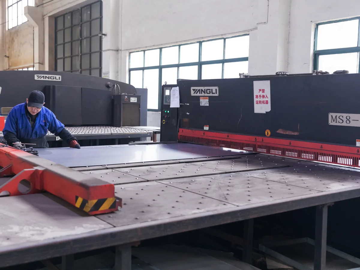 Keshun operator shearing steel sheet to size on the hydraulic guillotine beside the CNC turret punch
